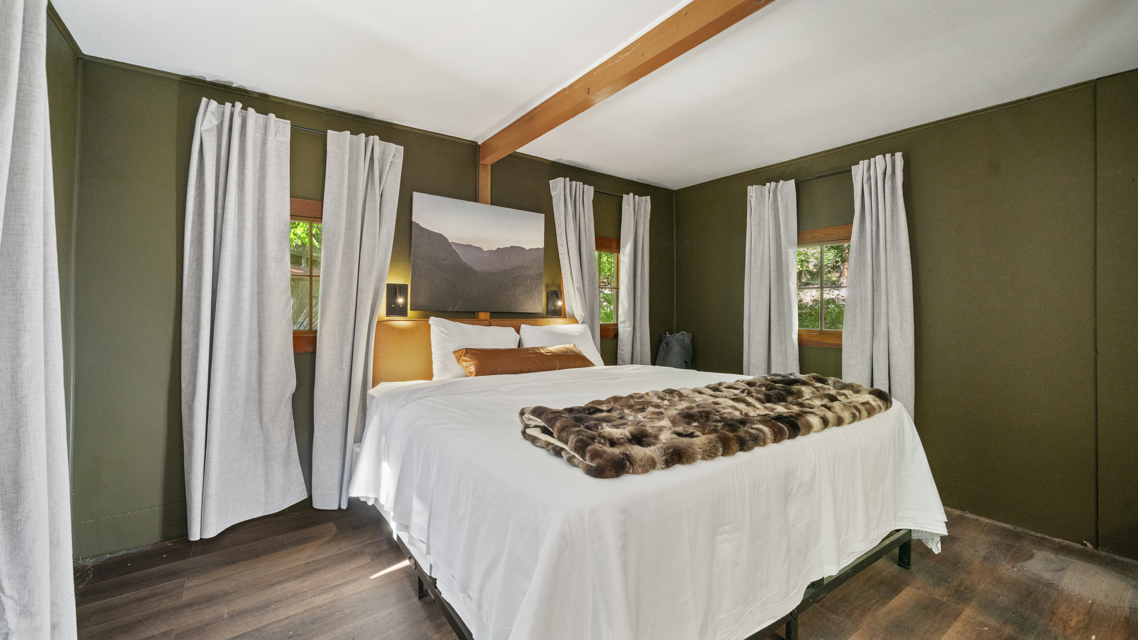 A wide-angle view of a bedroom with a large bed, white curtains, and artwork on the wall.