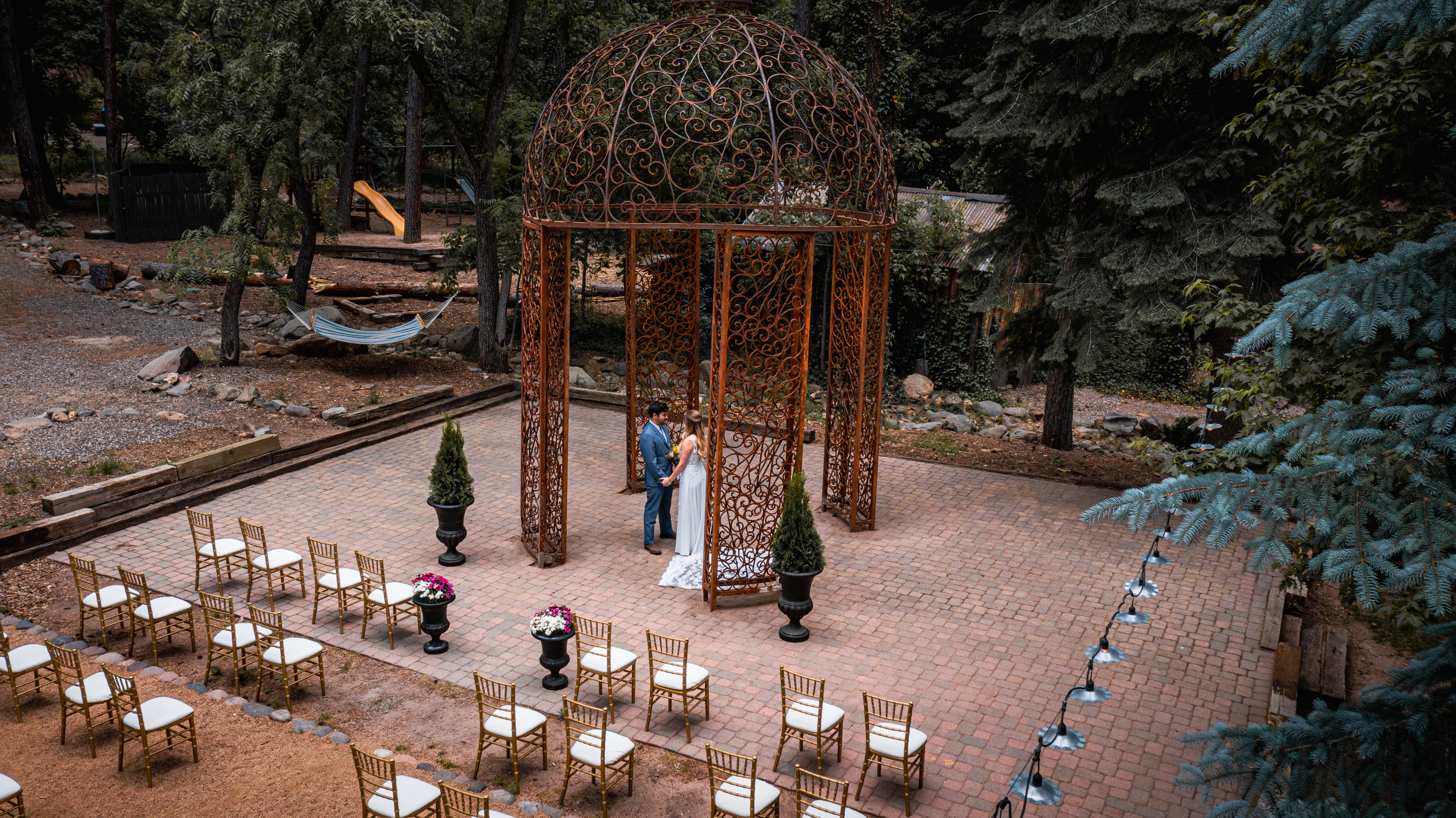 A beautifully set outdoor wedding scene with a gazebo, brick pavement, and surrounded by trees.