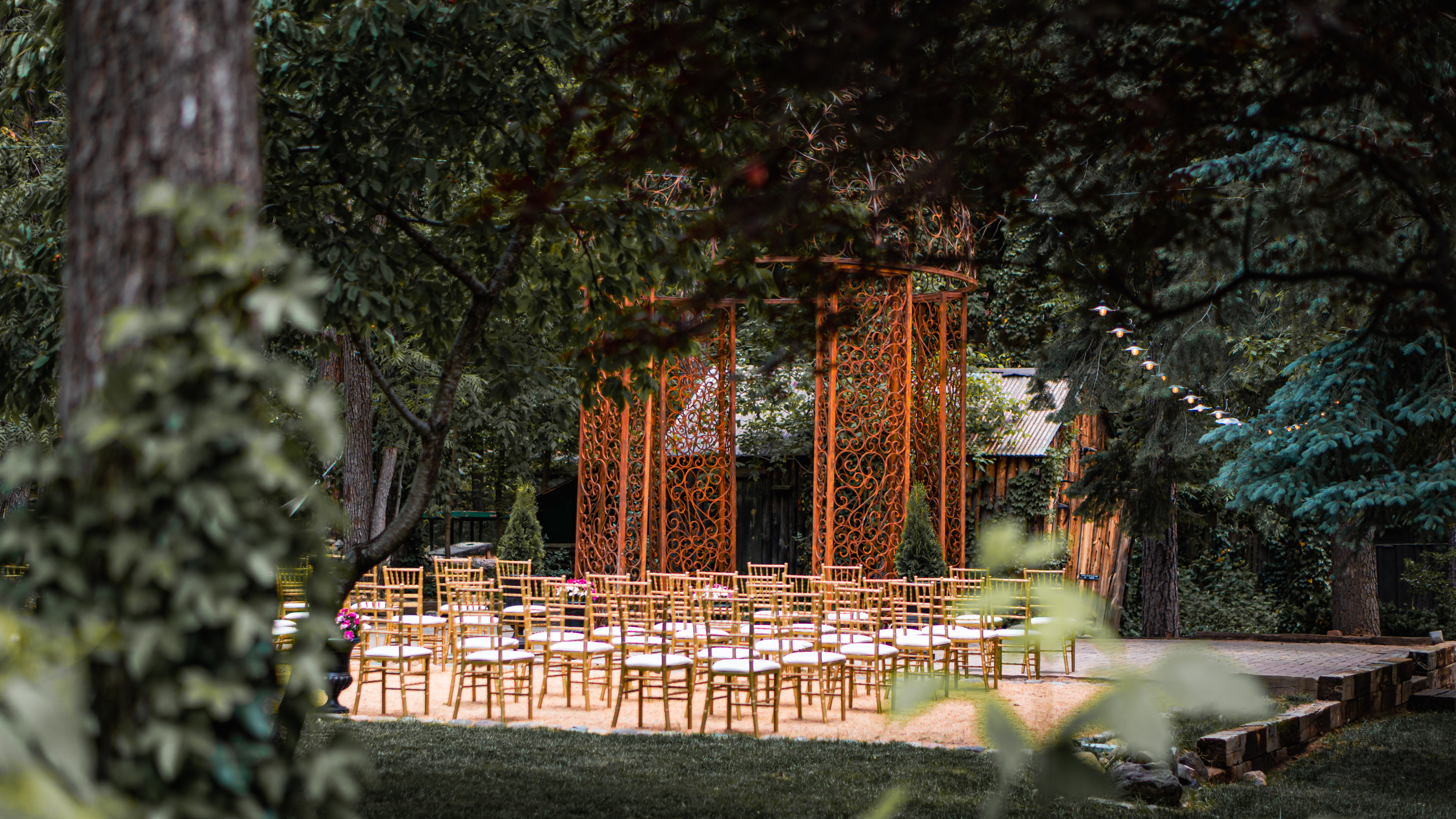 An outdoor event space with gold chairs arranged in rows, an orange metal archway, and surrounded by trees and grass.