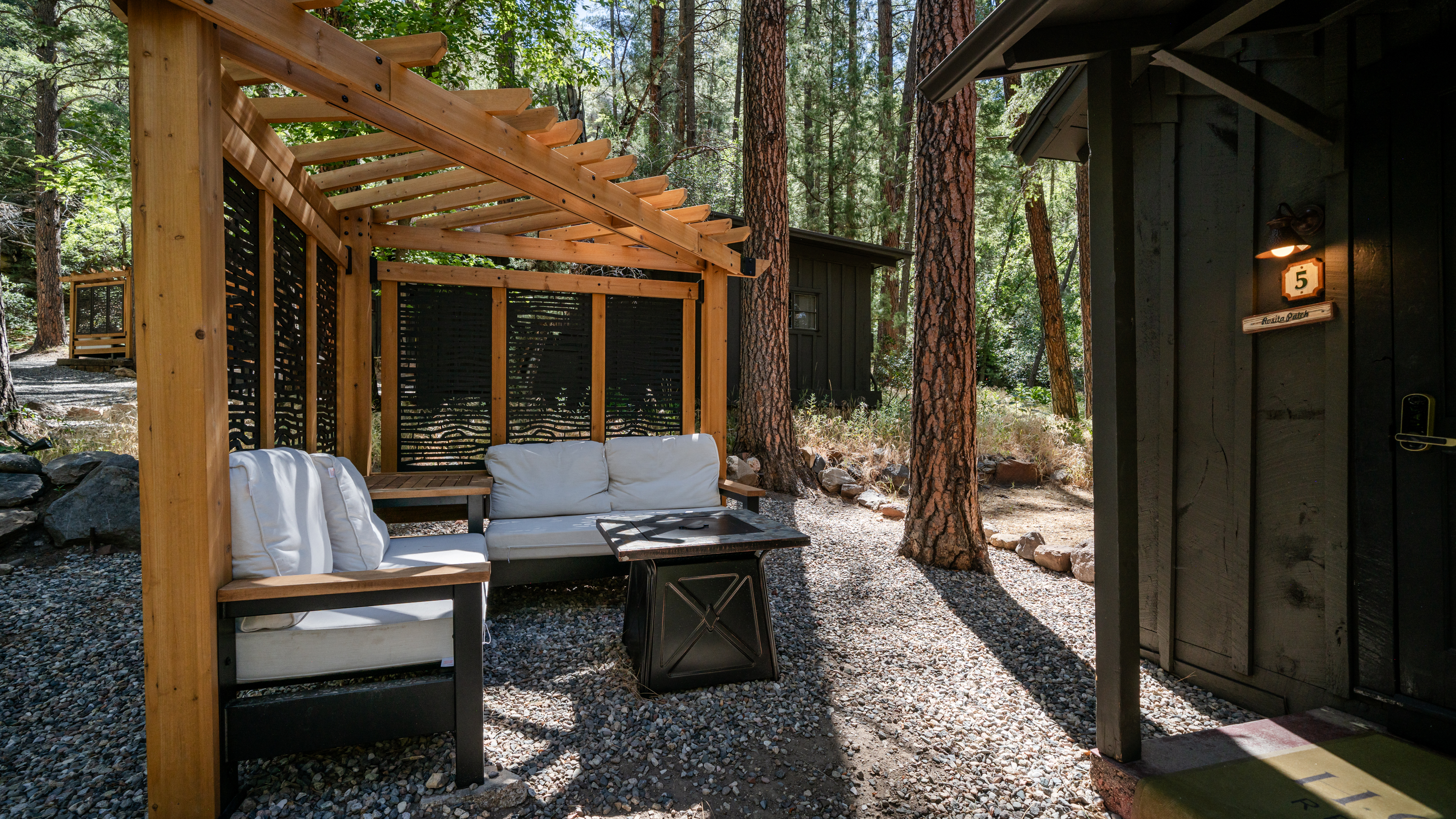 A seating area with a pergola, sofa, and table on gravel surrounded by trees.