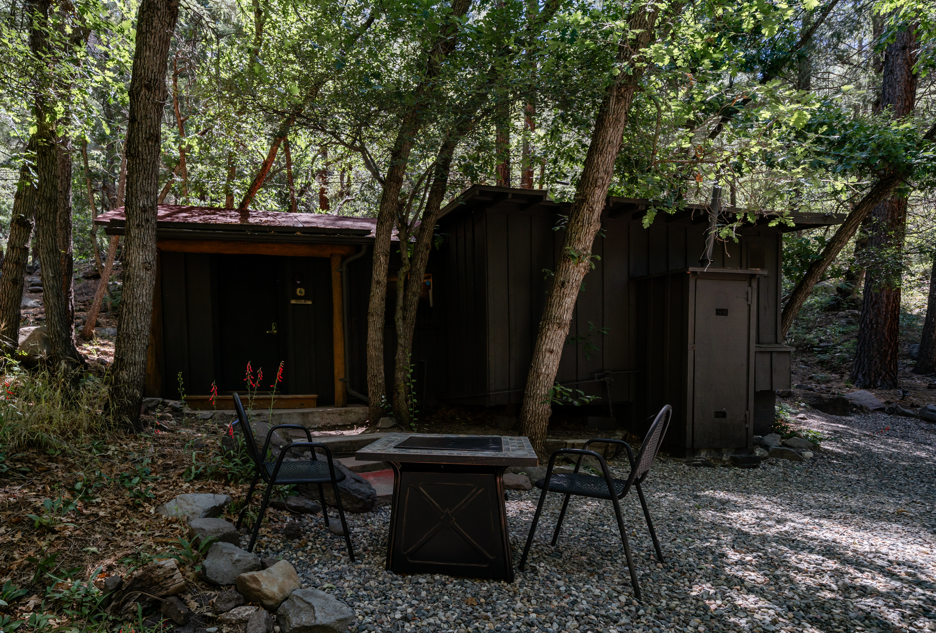 A serene outdoor seating area with a fire pit, table, and chairs surrounded by trees and a wooden building.