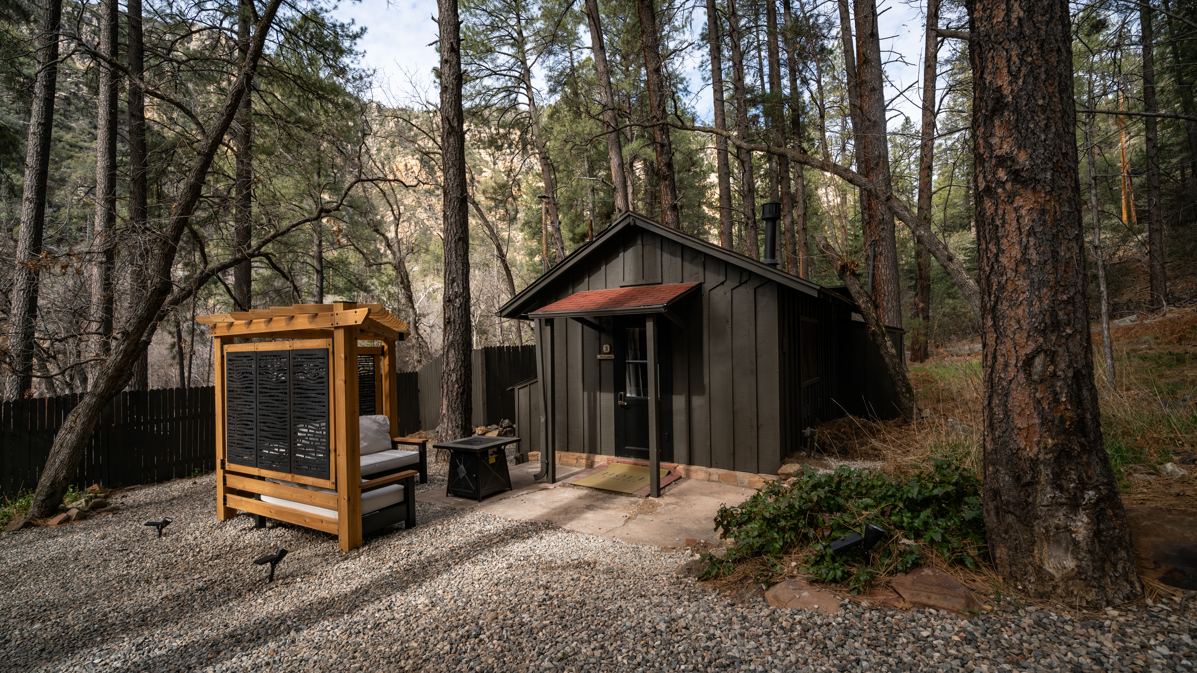A small wooden cabin with a red awning and a gravel driveway surrounded by trees.