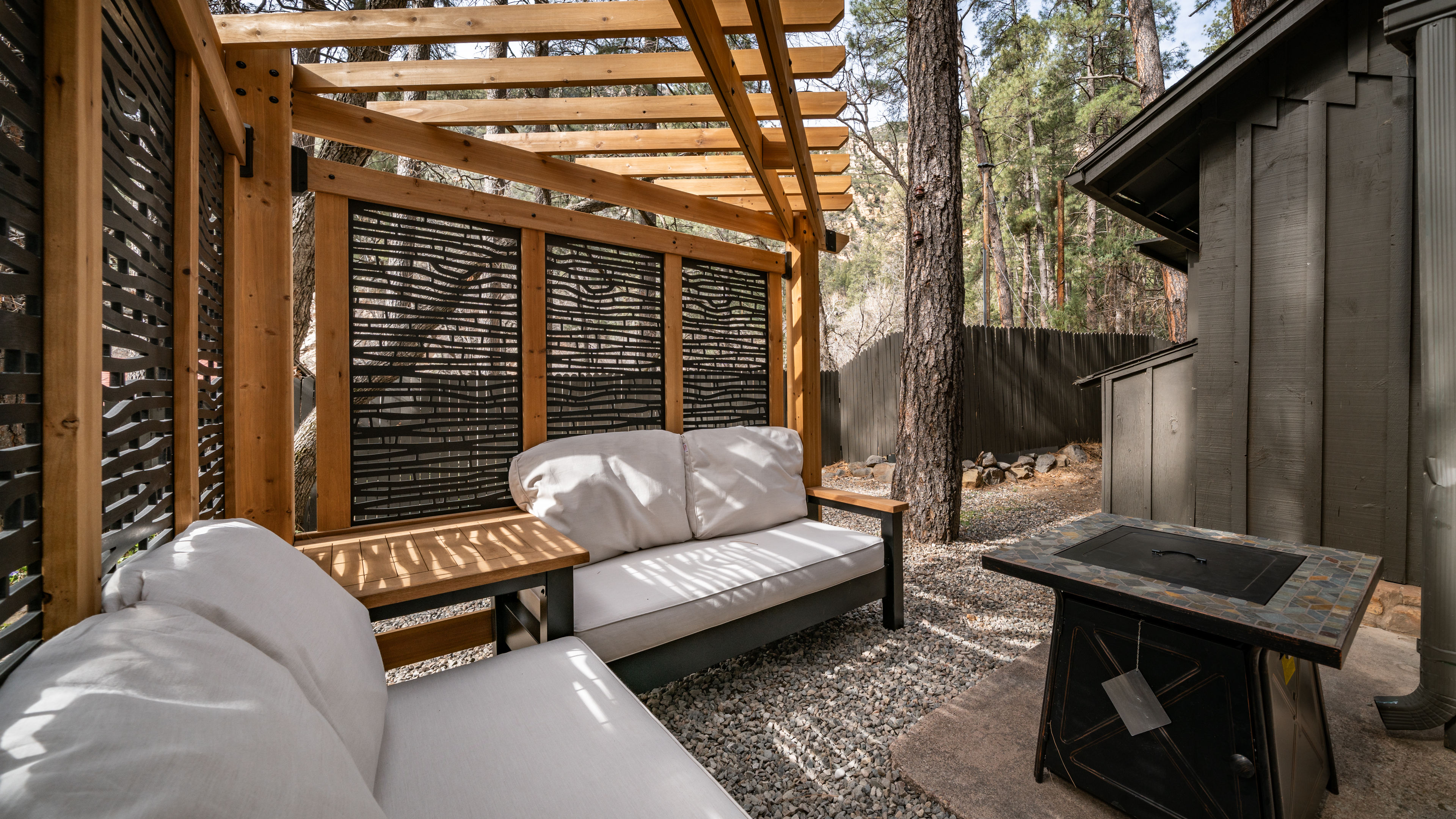 A cozy outdoor seating area with a wooden pergola, a white couch, and a tree in the background.